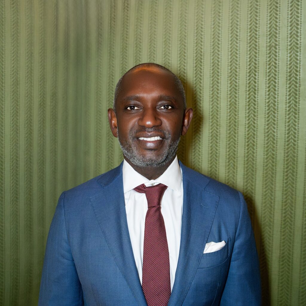 Co-founder Nick Jean-Baptiste, a man in a blue suit, white shirt, and patterned tie, smiles slightly.